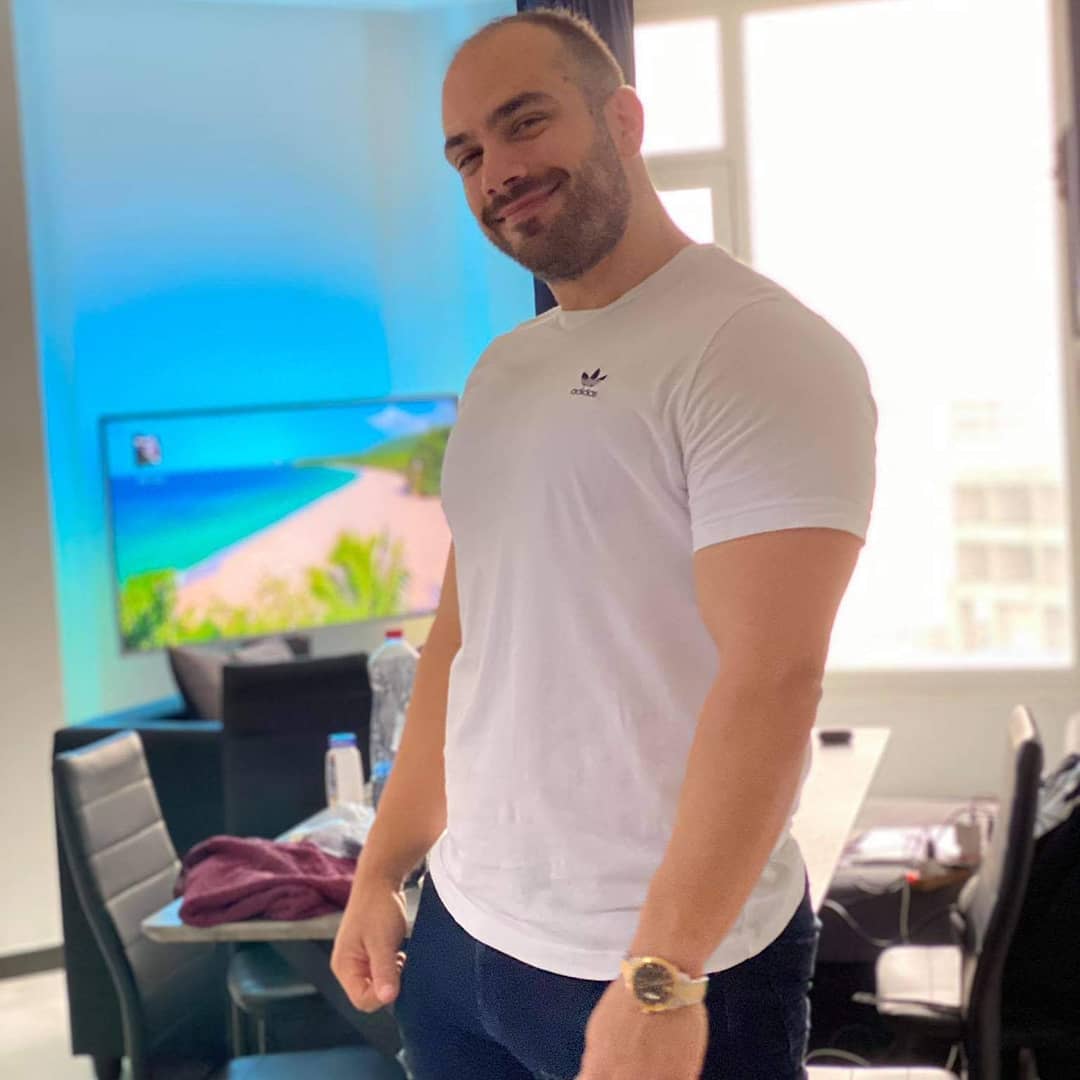 A man with a beard and a short haircut is standing and smiling in an office environment. He is wearing a white T-shirt with a small Adidas logo and dark pants. In the background, there are office chairs, a table with various items, and a large window with daylight streaming in. A television displays a tropical beach scene.